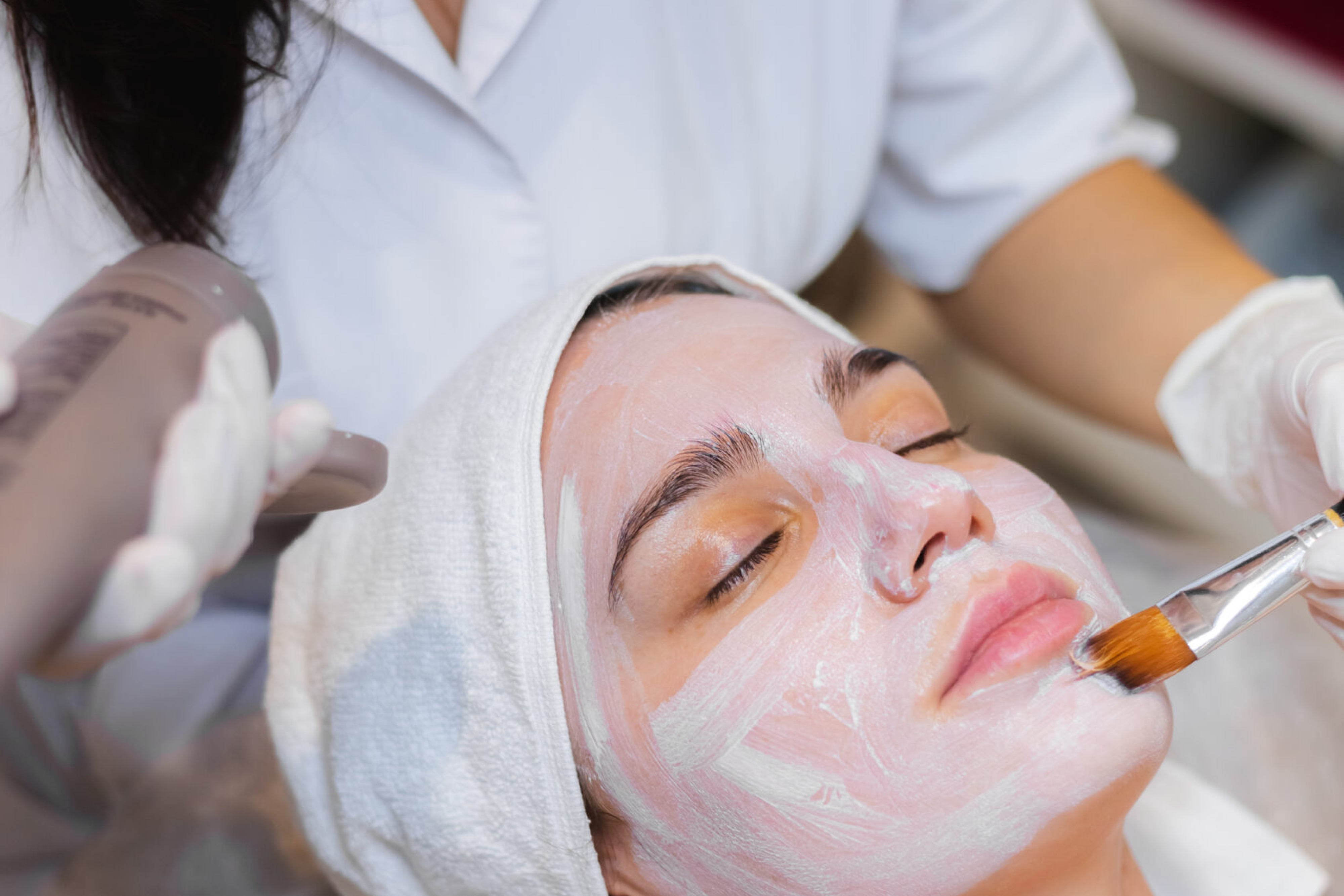 Beautician with a brush applies a white moisturizing mask to the face of a young girl client in a spa beauty salon